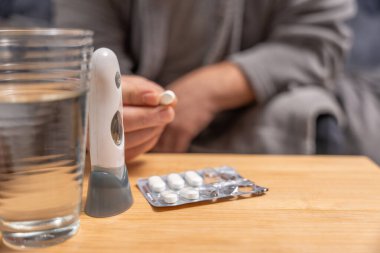 Yakın plan, termometre ve su bardağının yanında bir hap tutuyor. Alım, tedavi, sağlık ve hastalık yönetimi kavramı.