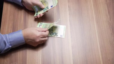 Masadaki Euro Banknotları sayılıyor. Yakın çekim görüntüsünde, ahşap bir masadaki 100 avroluk banknotları eller sayıyor. Olay yeri nakit işleme, finans ve günlük para işlemlerini gösteriyor.