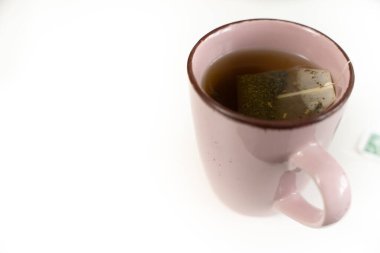 İçinde çay poşeti olan pembe bir kupa. Çay poşeti yeşil ve siyah. Kupa beyaz bir yüzeyde.