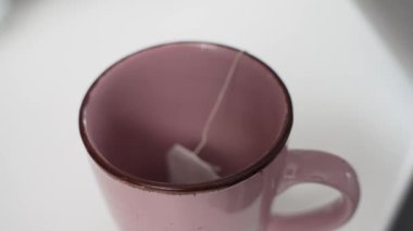 Sıcak su, çay poşetiyle pembe bir fincana dökülüyor. Nazik bir hareket ve renk dağılımı yaratıyor. Makro perspektif dokuyu, buharı ve demleme detaylarını vurguluyor.