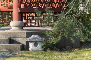 Bir bahçedeki geleneksel bir pavyonun yanında bir taş fener duruyor. Güneş ışığı, ahşap yapıları ve çevreleyen yeşillikleri aydınlatır.
