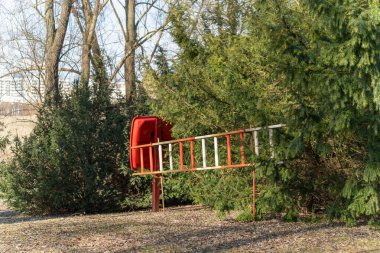 Parktaki ağaçların yanında parlak kırmızı bir kurtarma kutusu duruyor. Yanında bir merdiven var, acil durum için hazır..