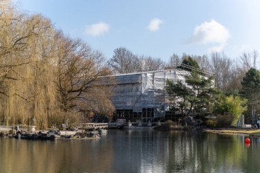 Sakin gölün yanındaki modern bina yapım aşamasında. Sahneyi sonbahar yapraklı ağaçlar oluşturuyor. Yansımalar suyun yüzeyinde yavaşça dalgalanır. İskeleli yapı doğal yeşilliğin yanında duruyor.