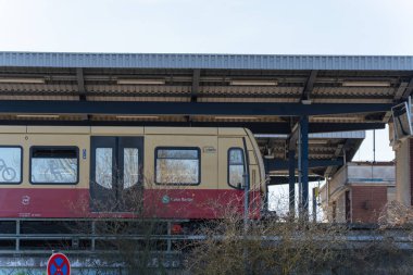 Metal istasyon tepe örtüsünün altında kırmızı ve krem rengi bir tren. Tepedeki yapıda yaş ve aşınma izleri var. Tren pencereleri solgun bir gökyüzünü yansıtır. Çalılıklar ve küçük bir işaret çerçevesi ön planda