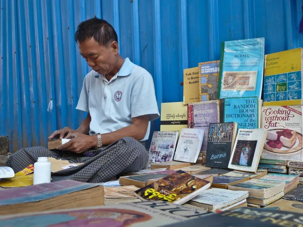 YANGON, MYANMAR - 23 DE DICIEMBRE DE 2013: Un librero callejero se sienta entre sus libros mientras hace una reparación —  Fotos de Stock