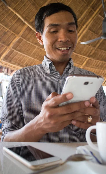 Low angle vertical medium shot of smiling male person of color looking at his smartphone 'Phablet' in a rustic beach cafe-like environment.