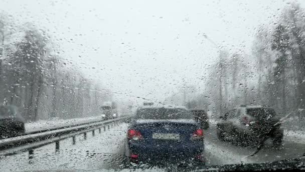 Une vue à travers le pare-brise d'une voiture avec des gouttes de neige fondue sur la route avec des voitures dans un embouteillage. — Video