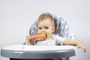 Beyaz bir arka planda yüksek bir sandalyede öğle yemeği için taze çiğ havuç yiyen şirin küçük çocuk. Yemekteki çocuklar için vitaminler.