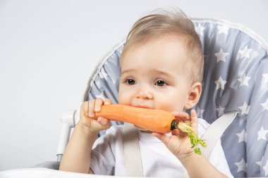 Beyaz bir arka planda yüksek bir sandalyede öğle yemeği için taze çiğ havuç yiyen şirin küçük çocuk. Yemekteki çocuklar için vitaminler.