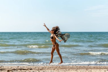 Aktif bir kadınla canlı deniz sahnesi, parlak deniz kıyısında süzülen akıcı elbiseli bir kadının büyüleyici görüntüsü.