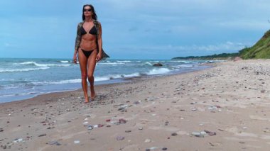 Siyah bikinili kadın kumlu sahilde kameraya doğru yürüyor, güneş gözlüklü güvenli adımlar, bacaklarının yakın plan detayları..