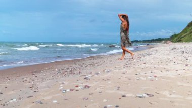 Uçan elbiseli kadın deniz kabuğu sahilinde yürüyor, esinti dalgalı saçlar, okyanus dalgaları ve çakıl taşları altında dalgın dalgın dalgın dalgın