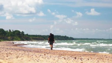 Sırt çantası ve sörf ile kumlu sahil şeridinde yürüyen erkek gezgin, dağınık bulutlu mavi gökyüzü, derin düşüncelere dalma hızı ve uzak mesafe