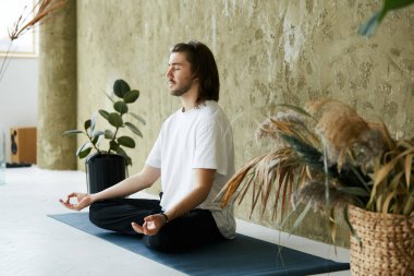 Rahat sakin bir adam modern tasarım evinde oturuyor ve meditasyon yapıyor. Yoga pozisyonunda ve konsantre olmuş sevimli bir erkek.