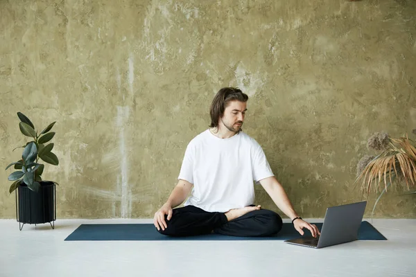 handsome man in lotus pose using laptop, male sitting on yoga mat and typing on computer in white shirt, copy space