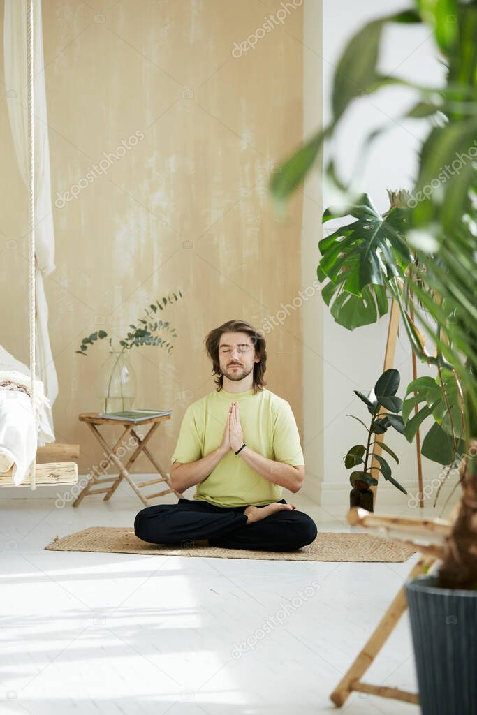 handsome man sitting in lotus pose holding arms in namaste, meditation teacher practicing concentration at home with green plants