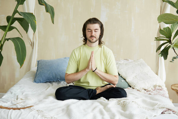 handsome man sitting on the bed in lotus pose and namaste, mindfulness and yoga concept, copy space