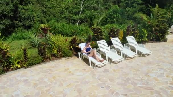 Girl in the outdoor lounge chair uses laptop for remote work on vacation — Wideo stockowe