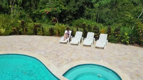 Aerial view of the girl laying in the outdoor lounge chair next to the pool. Uses laptop for remote work on vacation — Wideo stockowe