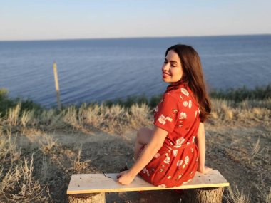 Kırmızılı kız bir bankta oturmuş güneşin doğuşunu seyrediyor.