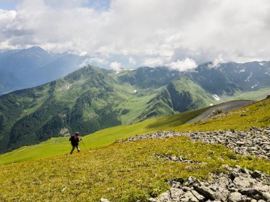 Sırt çantalı bir yürüyüşçü dağların arka planındaki çimenli yamaca gider. Arkadan bak. Arkhyz 'de güzel güneşli bir gün. Sağlıklı yaşam tarzı, yürüyüş aktivitesi, yürüyüş macerası kavramı