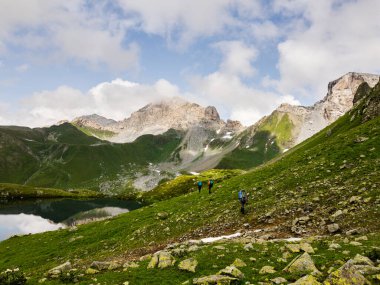 Kayaların üzerinde durup dağ gölüne bakan bir yürüyüşçü. Sırt çantalı bir adam şeffaf bir göle ve bulutlu dağlara karşı ayakta duruyor. Kafkas Dağları. Arkhyz. Rusya