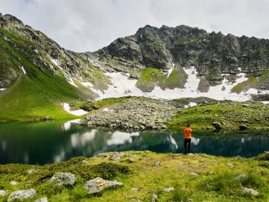 Kayaların üzerinde durup dağ gölüne bakan bir yürüyüşçü. Sırt çantalı bir adam şeffaf bir göle ve bulutlu dağlara karşı ayakta duruyor. Kafkas Dağları. Arkhyz. Rusya