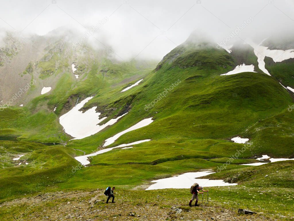 Hiker with a backpack goes on the grassy slope on a background of mountains in clouds. Concept of healthy lifestyle, trekking activity, hiking adventure