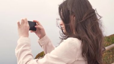Genç ve güzel bir kadın turist, Portekiz 'deki Cabo da Roca Burnu' nun batı kıyısında duruyor. Bulutlu bir gün. Kız akıllı telefonuyla okyanusu fotoğraflıyor. Saçlar uçuşuyor ve dalgalanıyor.