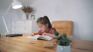 Masada oturan bir kız kitap okuyor. Çocuk okumayı öğreniyor. Evde eğitim. Akıllı bir çocuk. Kitap okuyorum. Çocuk masalları. Kendini soyutlama. Yavaş çekim