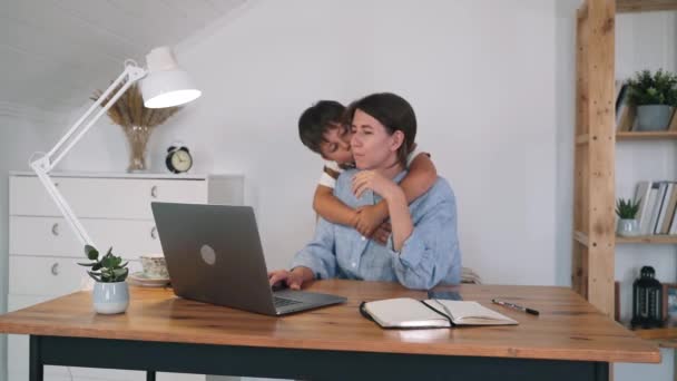 Uma mulher com um filho está a trabalhar num portátil. O menino abraça e beija sua mãe enquanto trabalha. Uma mulher beija o filho. Apoio na família. Freelance. Auto-isolamento. Trabalho em casa — Vídeo de Stock