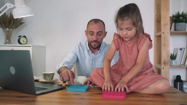 Un uomo e un bambino stanno giocando a Poppit. Un uomo che lavora in un computer portatile si prende una pausa e gioca Poppit con una ragazza. Un giocattolo anti-stress. Tendenze popolari del 2021. Auto-isolamento. Rilassati — Video Stock