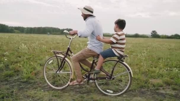 Un hombre lleva a un niño en una bicicleta retro. Padre e hijo están conduciendo por un campo de flores. Estilo retro. Una bicicleta de moda. Autoaislamiento. Recreo al aire libre. Vida en el campo cámara lenta — Vídeo de stock