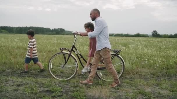 Um homem com filhos anda de bicicleta retro. Uma família com uma bicicleta está descansando em um campo de flores. Estilo retrô. Uma bicicleta na moda. Auto-isolamento. Férias em família. Paternidade. Vida no campo Movimento lento — Vídeo de Stock