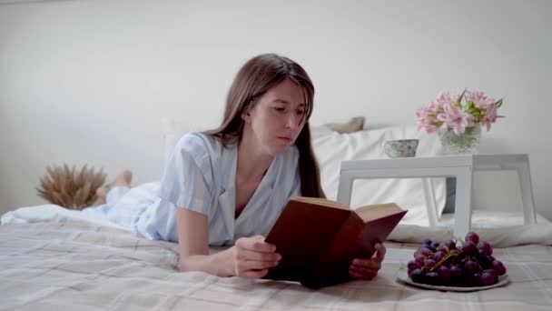 A woman reads a book while lying in bed. A woman dreams. A bedside table with flowers and a cup of coffee. Rest at home. Reflections. Beautiful pajamas on a woman. Bedding. Rug. Cozy bed. Slow motion — Stock Video