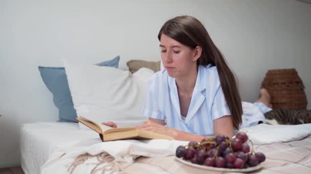 Una donna legge un libro sdraiata a letto Una donna sfoglia una pagina di Uva su un piatto Un gatto addormentato Riposo a casa. Bellissimo pigiama su una donna. Biancheria da letto. Tappeto. Letto accogliente. Rallentatore — Video Stock