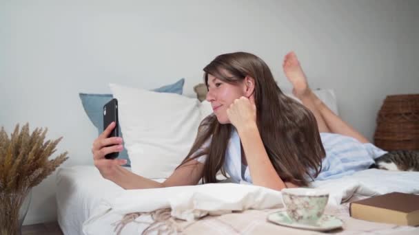 Vrouw aan de telefoon online liggend in bed Vrouw lachend Slapende kat Ontspannend thuis. Mooie pyjama op een vrouw. Een kopje koffie Beddengoed Een boeket gedroogde bloemen Een gezellig bed. Langzame beweging — Stockvideo