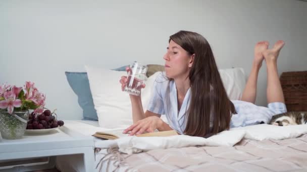 Жінка читає книгу, що лежить у ліжку Жінка п'є воду Приліжковий стіл з квітами та склянкою води Розслабляючись вдома Красива піжама на жінці Постіль. Рег. Затишне ліжко. Повільний рух — стокове відео