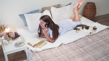 Bir kadın yatakta uzanıp telefonda konuşuyor. Yatakta kahvaltı yapıyor. Üzerinde çiçekler ve bir fincan kahve olan bir komodin. Evde dinlenin. Bir kadında güzel pijama. Yatak çarşafları. Sıcak yatak. Yavaş çekim
