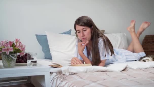 En kvinna som läser en bok liggande i sängen En kvinna som rätar ut håret Ett nattduksbord med blommor och ett glas vatten Slappna av hemma Vacker pyjamas på en kvinna Sängkläder. Mugg. Mysig säng. Långsamma rörelser — Stockvideo