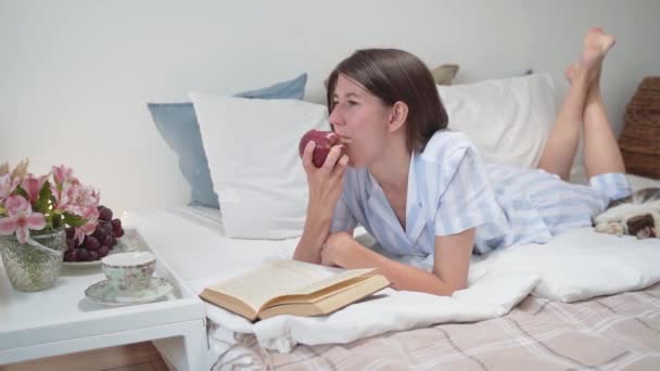 Μια γυναίκα τρώει ένα μήλο και διαβάζει ένα βιβλίο ξαπλωμένη στο κρεβάτι. Πρωινό στο κρεβάτι. Ένα κομοδίνο με λουλούδια και ένα φλιτζάνι καφέ. Χαλαρώστε στο σπίτι. Ωραίες πιτζάμες σε μια γυναίκα. Άνετο κρεβάτι. Αργή κίνηση — Αρχείο Βίντεο