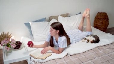 Bir kadın yatakta yatarken bir elma yer ve bir kitap okur. Yatakta kahvaltı. Üzerinde çiçekler ve bir fincan kahve olan bir komodin. Evinde dinlen. Bir kadında güzel pijama. Sıcak yatak. Yavaş çekim