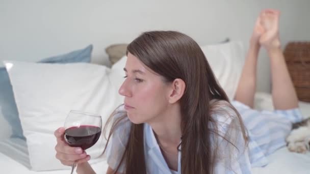 Uma mulher bebe vinho deitada na cama. Uma mulher segura um copo de vinho. Relaxe em casa. Belo pijama numa mulher. Roupa de cama. Cama acolhedora. Movimento lento. Fechar — Vídeo de Stock