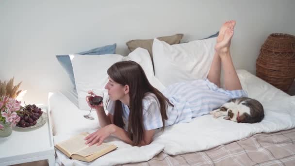 一个女人躺在床上看书，喝酒。在床上吃晚餐床头柜上放着花和葡萄.一杯葡萄酒。在家里放轻松女人穿上漂亮的睡衣舒服的床慢动作 — 图库视频影像