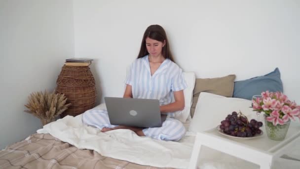 Une femme travaille sur un ordinateur portable alors qu'elle est assise au lit Dîner au lit. Table de chevet avec fleurs et raisins. Un verre de vin. Travailler à la maison. Beau pyjama sur une femme. Un chat endormi. Lit douillet. Mouvement lent — Video