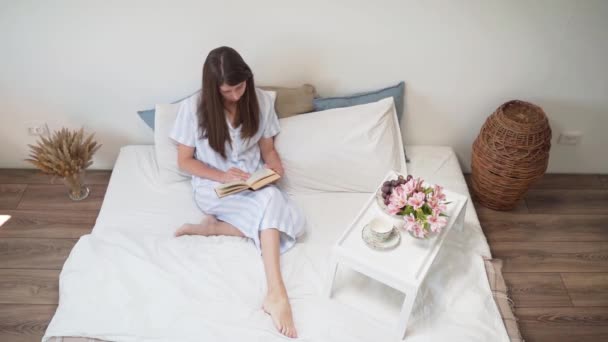 Yatakta oturan bir kadın kitap okuyor. Üzerinde çiçekler ve bir fincan kahve olan bir komodin. Evinde dinlen. Bir kadında güzel pijama. Yatak örtüsü. Sıcak yatak. Bir buket kurumuş çiçek. Yavaş çekim — Stok video