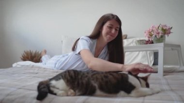 Bir kadın yatakta yatarken bir kediyi okşar. Evde dinlenin. Çiçek ve üzümlü komodin. Bir kadında güzel pijama. Hayvan bakımı. Yatak çarşafları. Halı. Sıcak yatak. İç mekan. Kahvaltı Ağır çekim