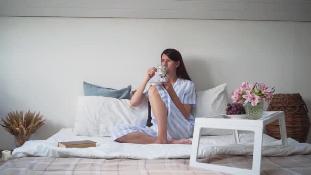 Una mujer bebe café sentada en la cama. Mesita de noche con flores y uvas. Relájate en casa. Hermoso pijama para una mujer. Ropa de cama. Acogedora cama. Un ramo de flores secas. Movimiento lento — Vídeo de stock