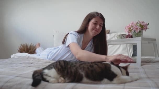 Uma mulher acaricia um gato enquanto estava deitada na cama. Descanse em casa. Mesa de cabeceira com flores e uvas. Belo pijama numa mulher. Cuidados com animais. Roupa de cama. Tapete. Cama acolhedora. Interior. Pequeno-almoço câmera lenta — Vídeo de Stock