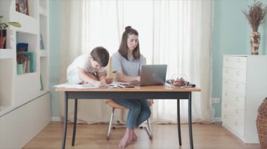 Annem evde, masada internet üzerinden çalışıyor. Masada oturan küçük bir çocuk. Oğlu çizimini gösteriyor. Uzaktan kumanda. Aile bireysel izolasyonda. Merkez ofis. Mutlu bir çocukluk. Karantinaya alın. Salgın.